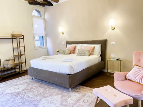Luxurious Verona suite with gray upholstered bed, white linens, pink accents, wooden beams, arched window, and elegant