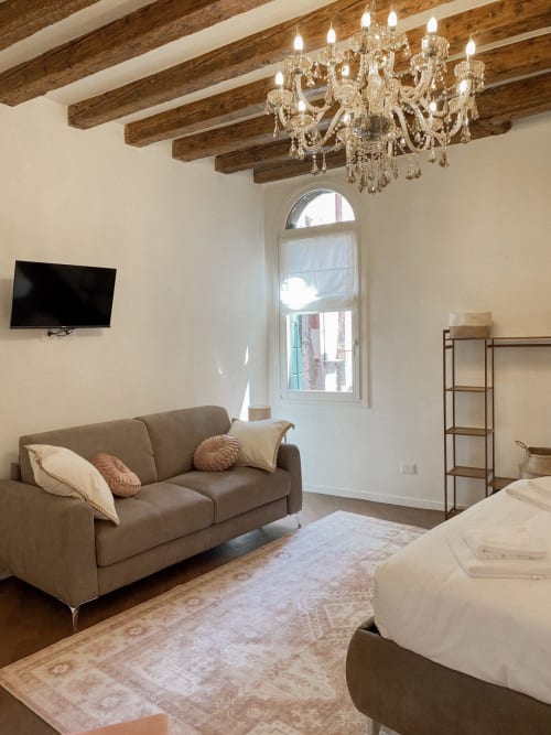 Luxurious suite in Verona with crystal chandelier, exposed beams, taupe sofa, flat-screen TV, and arched window with river