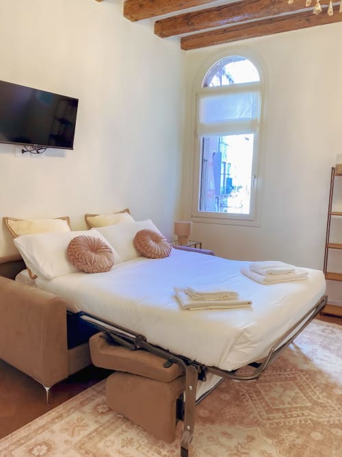 Bright suite bedroom with sofa bed, decorative pillows, exposed beams, arched window with canal view in Verona
