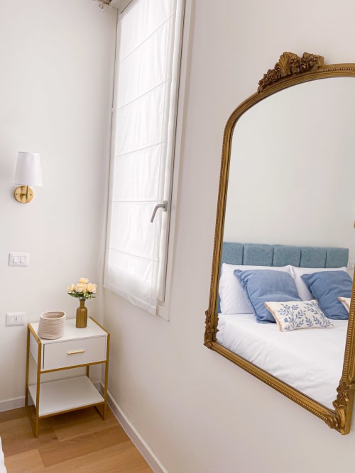 Luxury bedroom in Verona with ornate gold mirror, blue headboard, white nightstand, and natural light
