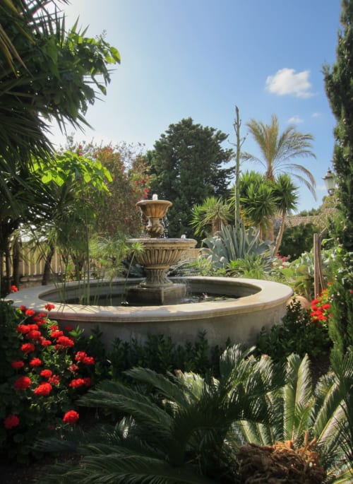 Lussureggiante giardino interno dell'Hotel Candalú con fontana ornamentale, fiori rossi, palme e vialetti in pietra a Valderice
