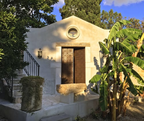 Cappella villa Valderice con facciata in pietra bianca, croce, portali in legno e vista del rigoglioso giardino dell'Hotel Candalú
