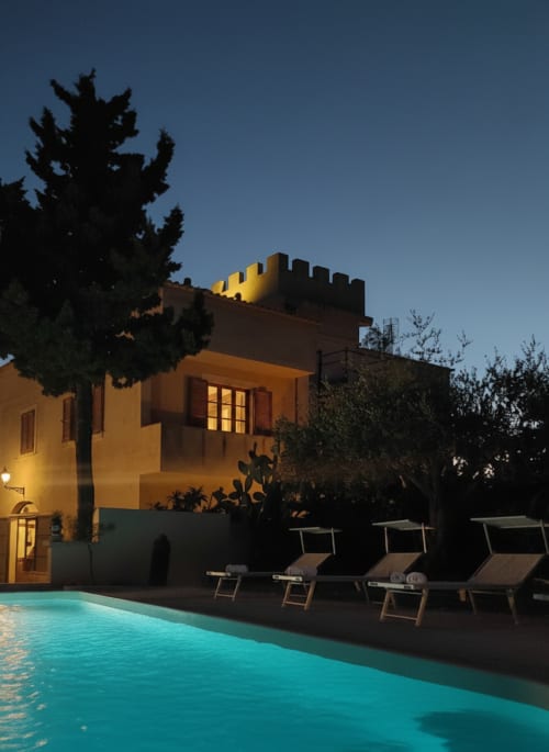 Suite dell'Hotel Candalú al tramonto con torre in stile castello illuminata, piscina turchese, lettini e vista sul giardino