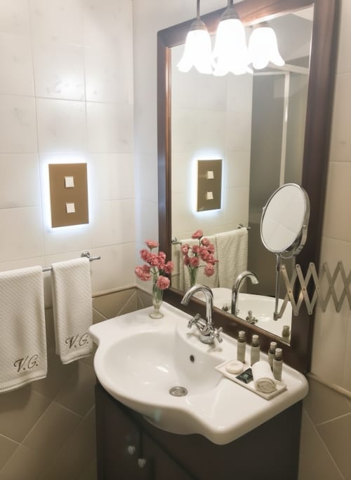 Bagno moderno con lavandino bianco, specchio illuminato a tre luci e fiori rosa nella Suite Interne dell'Hotel Candalú