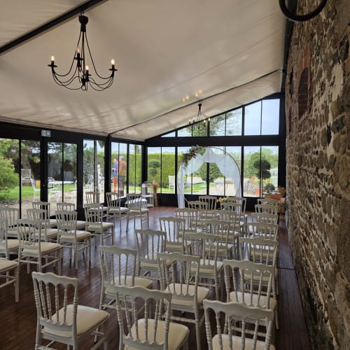 Elegant reception hall at Manoir de la Bégaudière with white chairs, stone walls, chandeliers, and garden views through large