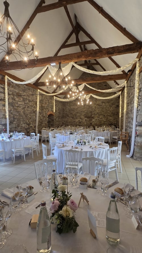 Elegant banquet hall at Manoir de la Bégaudière with exposed beams, draped lighting, white chairs, and stone walls set for