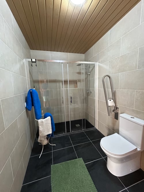 Banheiro moderno e acessível na casa de hóspedes Casa das Rodas com piso de azulejos pretos, paredes bege, teto de madeira e chuveiro com vidro