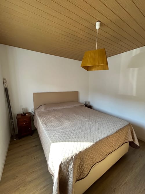 Quarto luminoso da pousada Casas do Sinhel com cama queen, cabeceira bege, teto de madeira, luminária amarela e paredes brancas