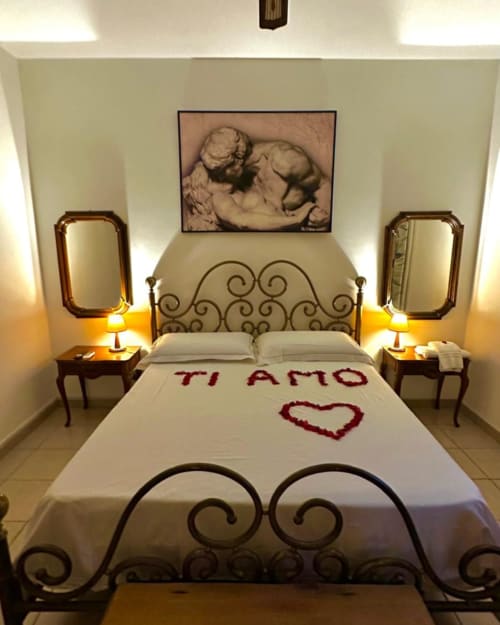 Camera romantica con letto in ferro battuto ornamentale, petali di rose 'Ti Amo', specchi, lampade e opere d'arte con cherubini a Pupi Catania B&B
