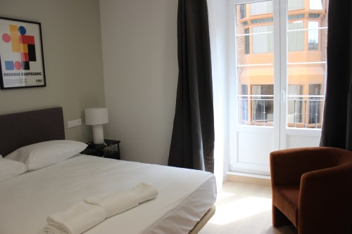 Moderna habitación de hotel en Palacio Mambrilla Valladolid con balcón con vista a la ciudad, cama blanca y butaca marrón