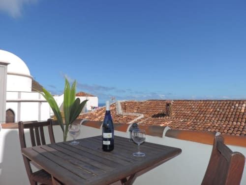 Terraza en la azotea de Casa Rural Los Helechos con botella de vino, copas y vistas al océano sobre edificios tradicionales con tejados de barro