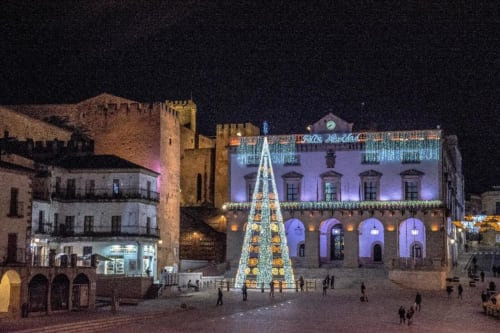 Plaza Mayor de Cáceres iluminada navideñamente con árbol decorado, edificios históricos y arquitectura medieval de noche