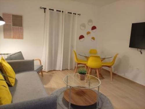 Salón moderno con sillas amarillas, sofá gris, mesa de comedor y televisor en Cáceres