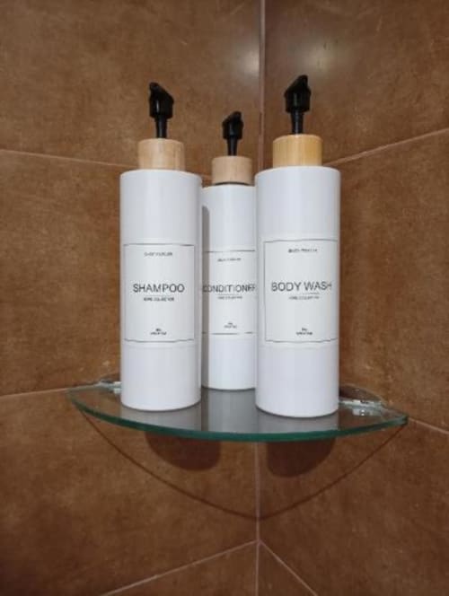 Tres botellas blancas de amenidades de baño con tapas de madera en un estante de vidrio del baño con azulejos en Apartamentos La Machacona.