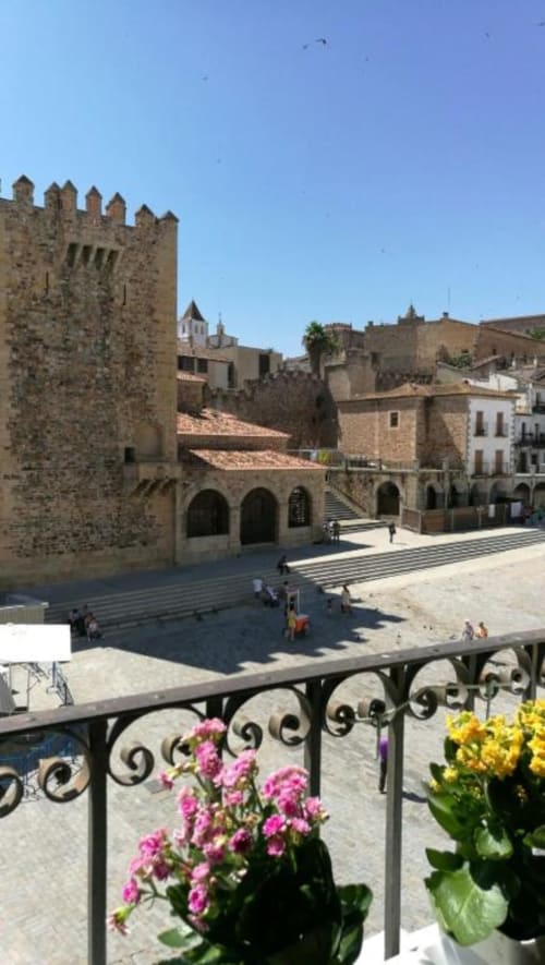 Plaza Mayor histórica con torre del castillo, arquitectura de piedra y flores rosas en la barandilla del balcón en Cáceres, España