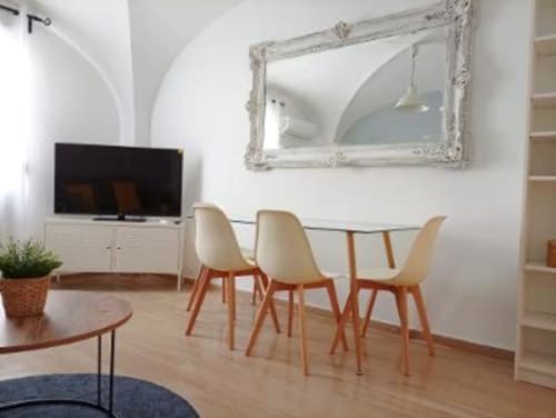 Comedor moderno en apartamento de Cáceres con mesa de cristal, sillas color crema, TV y espejo decorativo con vistas a Plaza Mayor