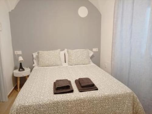 Dormitorio moderno en apartamento de Cáceres con cabecero gris, ropa de cama beige, dos toallas marrones, almohadas blancas