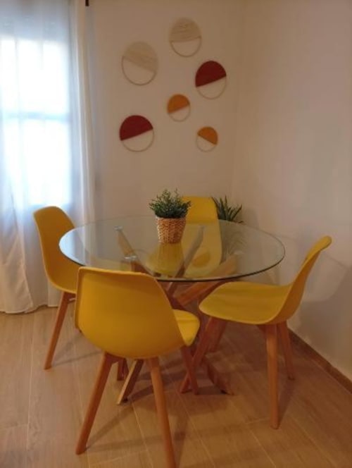 Comedor moderno con sillas amarillas y mesa redonda de cristal, plantas en macetas y arte mural colorido en apartamento de Cáceres