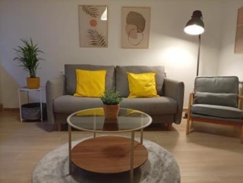 Moderna sala de estar en apartamento de Cáceres con sofá gris, cojines amarillos, mesa de cristal y decoración botánica