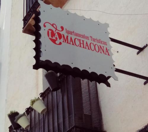Letrero de Apartamentos Turísticos La Machacona en Cáceres, con letras rojas sobre fondo blanco y bordes decorativos oscuros
