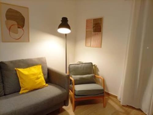 Zona de estar moderna en Cáceres con sofá gris, cojín amarillo, sillón de madera y lámpara de pie