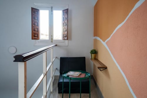 Rellano de escalera en apartamento de Cáceres con arte geométrico en pared, ventana, barandilla y mesita verde con revistas
