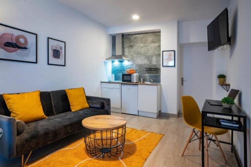 Moderno apartamento de 1 dormitorio en Cáceres con sofá negro, detalles amarillos, cocina americana y TV de pantalla plana