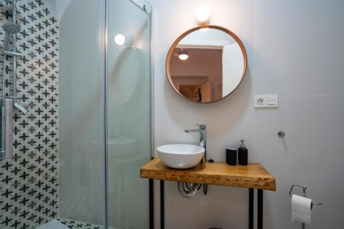 Baño moderno con espejo redondo de cobre, lavamanos blanco sobre vanidad de madera, accesorios negros y ducha estampada