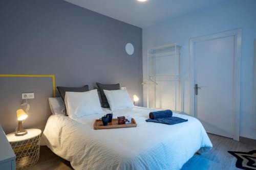Dormitorio moderno en Apartamentos La Machacona de Cáceres con cama blanca, pared gris destacada, baño privado y toallas azules.