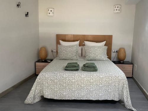 Dormitorio moderno en apartamento de Cáceres con cabecero de madera, ropa de cama estampada, toallas verdes, lámparas de mimbre y paredes blancas.