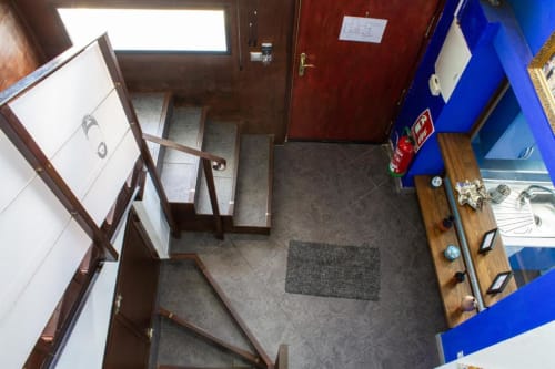 Entrada de apartamento bajo en Cáceres con escaleras, suelo de baldosa oscura, barandillas de madera y pared azul con cocina