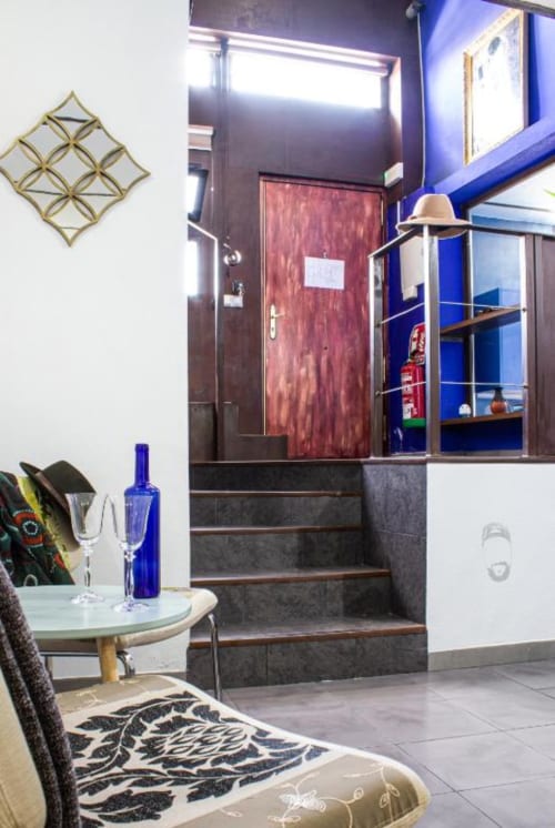Entrada moderna de apartamento en Cáceres con escaleras, pared azul, puerta de madera, mesa redonda con copas de vino y detalles geométricos