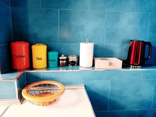 Estante de cocina en apartamento de Cáceres con botes de café, té, azúcar y tetera roja sobre pared de azulejos azules
