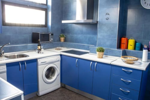 Cocina moderna azul con encimeras blancas, lavadora y electrodomésticos de acero inoxidable en apartamento de Cáceres