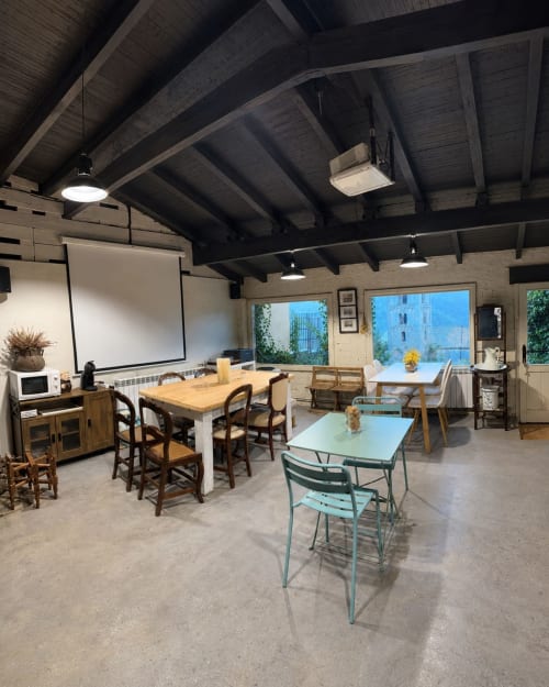 Sala común de Casa Tapiolas con mesas de madera, sillas, pantalla de proyección y vistas a las montañas en Molló, Girona