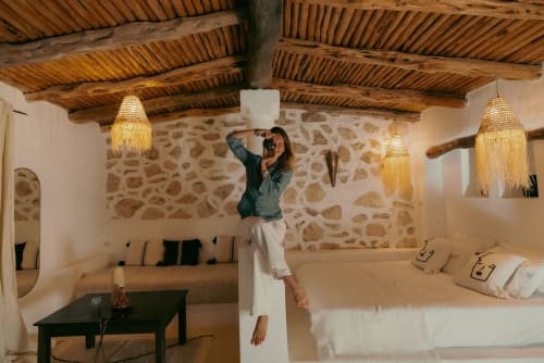 Femme ajustant un luminaire au plafond dans une chambre en pierre brute à l'hôtel Eucalyptus Ethnic d'Essaouira, avec poutres en bois et décor traditionnel