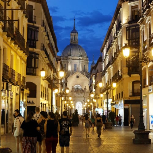 Calle peatonal al atardecer en Zaragoza con basílica histórica de cúpulas iluminadas, edificios resplandecientes y tiendas
