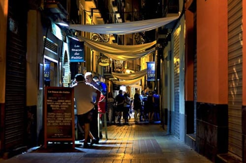 Estrecho callejón peatonal nocturno en el Centro de Zaragoza con iluminación dorada, visitantes y tiendas incluyendo Hostal