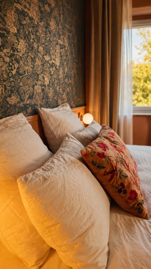 Chambre accueillante à La Maison 1903 avec papier peint à motifs, oreillers crème, coussin fleuri et fenêtre sur la verdure