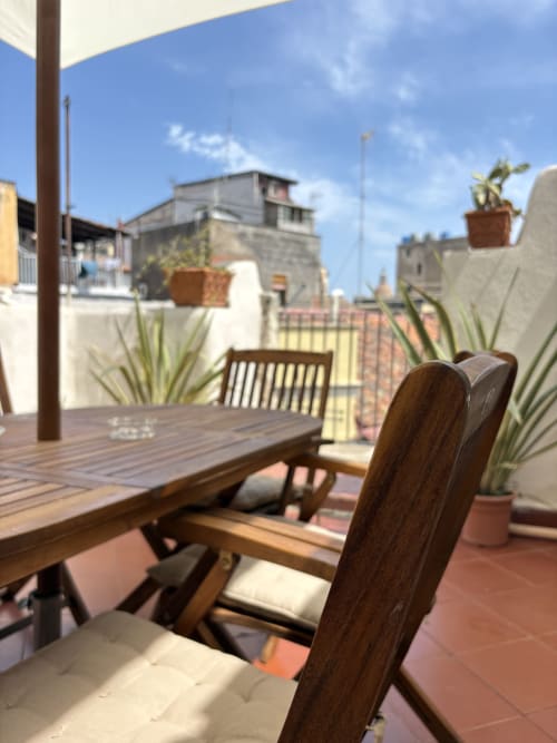 Tavolo e sedie in legno su una terrazza soleggiata con piante in vaso, con vista sullo skyline di Catania e gli edifici storici