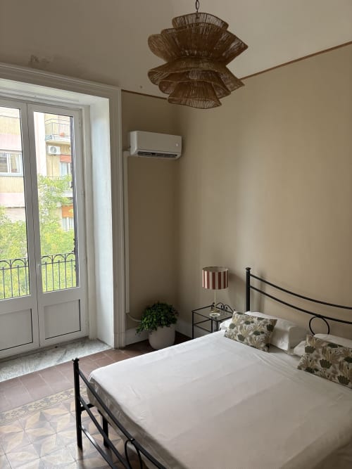 Camera luminosa a Sicily In Home Catania con letto matrimoniale, lampada a soffitto in rattan, balcone con vista città, aria condizionata e