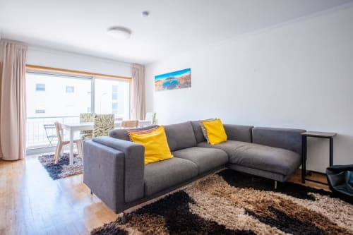 Modernes Wohnzimmer mit grauem L-förmigem Sofa, gelben Kissen, Holzböden und Balkon mit Blick auf Arcozelo