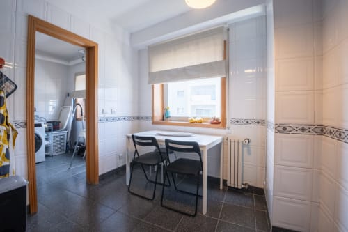 Helles Esszimmer mit weißen Fliesen, Holzfensterrahmen, zwei schwarze Stühle am weißen Tisch und Heizkörper im EcoHost-Apartment