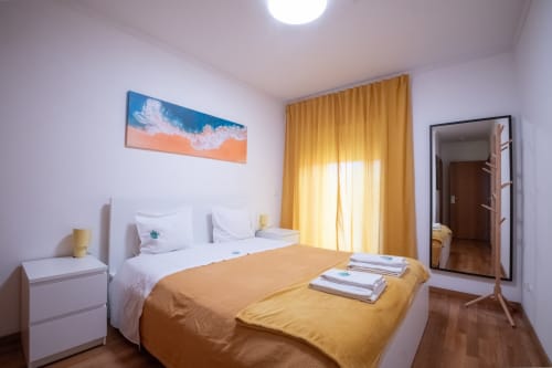 Helles Schlafzimmer im Gaia Beach&Golf mit gelben Vorhängen, Doppelbett, weißem Nachttisch und Strandmotiven