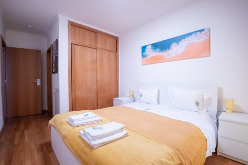 Helles Schlafzimmer im EcoHost Gaia Beach&Golf mit gelbem Überwurf, weißer Bettwäsche, Holzschrank, Strandkunst und Holzdetails