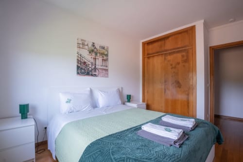 Helles Schlafzimmer im EcoHost Arcozelo mit Queensize-Bett, weißer Bettwäsche, grünen Akzenten, Holzkleiderschrank und Fahrrad-Kunstwerk