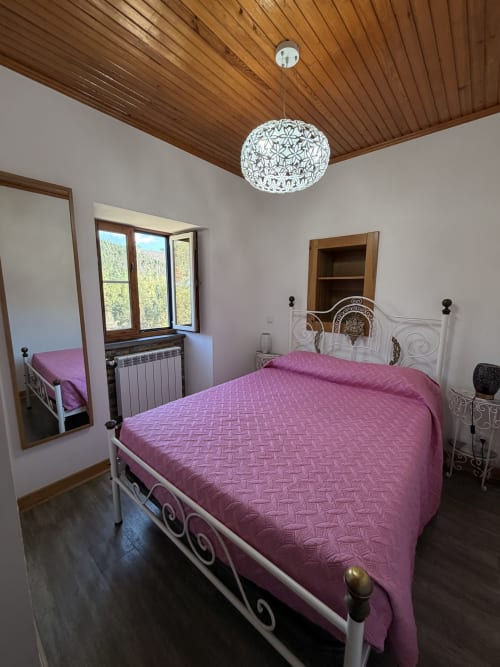 Quarto moderno com roupa de cama rosa, cama de ferro forjado, teto de madeira, luminária suspensa branca e vista para as montanhas pela janela