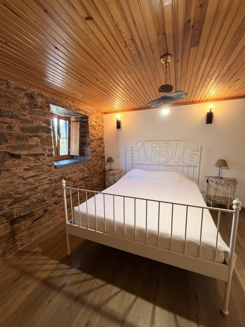 Quarto rústico na casa da Adega em Casas do Sinhel com teto de madeira, paredes de pedra, cama branca de ferro e janela.