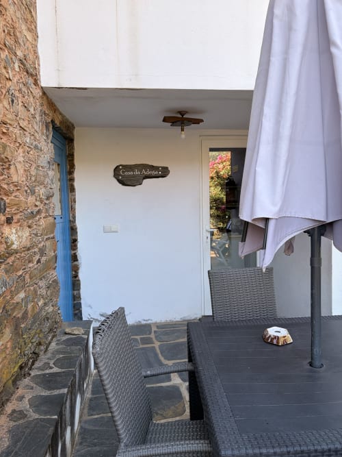 Entrada da casa rural Casa da Adega em Casas do Sinhel, com muros de pedra, porta azul, pátio com assentos e chapéu-de-sol em Relva da Mó