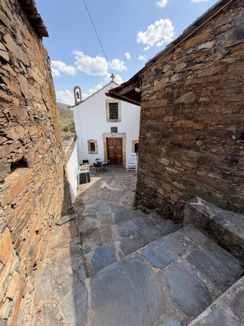 Caminho de pedra entre muros rústicos levando à Casa da Capela, uma vila de estilo capelinha com paredes brancas em Roda Fundeira com sino